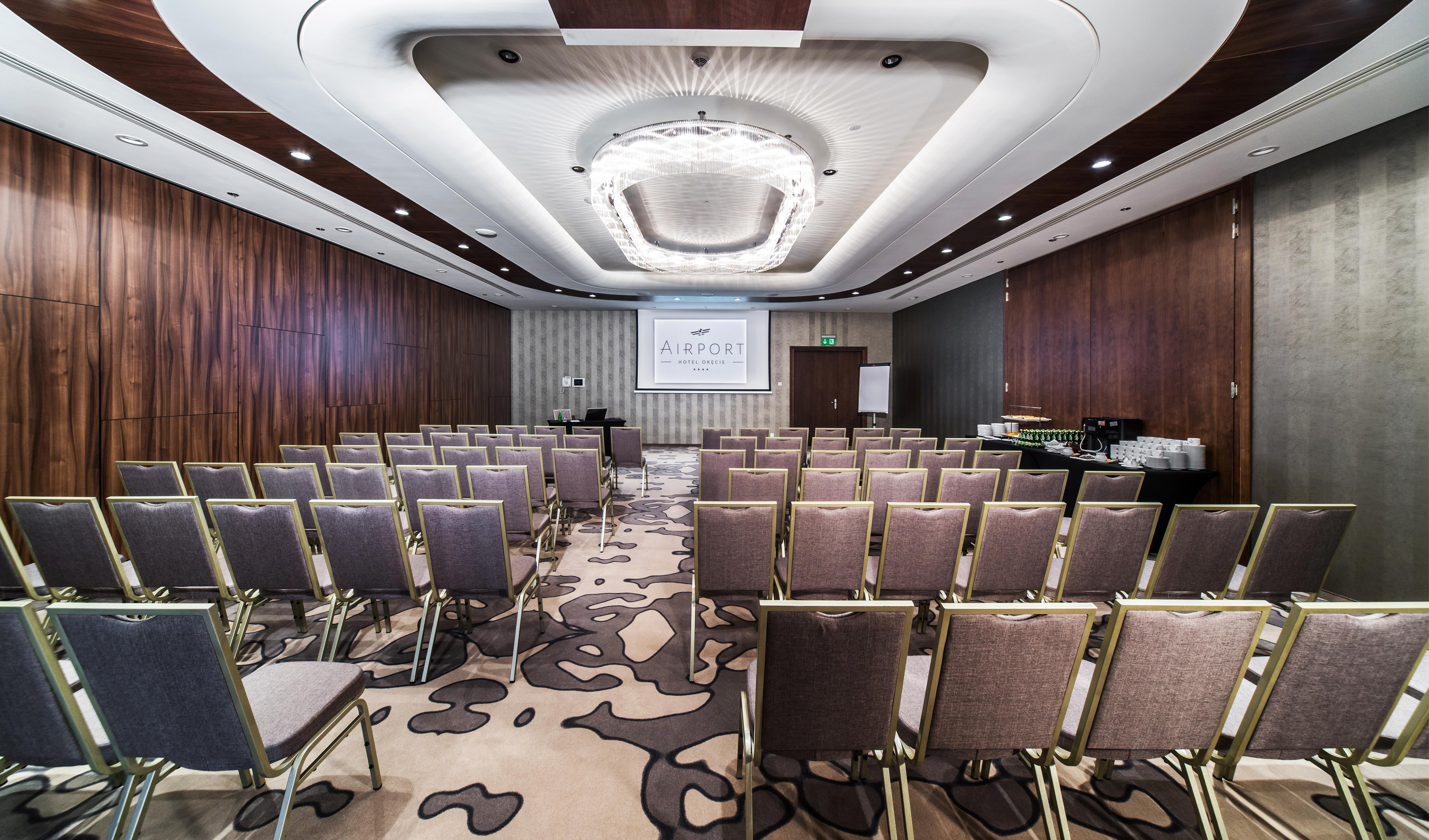 Airport Hotel Okęcie Warschau Buitenkant foto The photo shows a conference or meeting room set up for an event. It features rows of gray chairs arranged facing a presentation area with a screen. The walls are designed with a combination of wood paneling and light-colored wallpaper. A modern chan